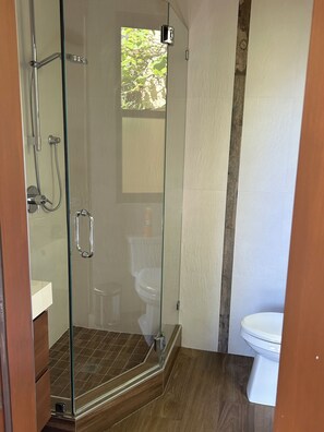 Une douche en verre avec un sol en bois, des toilettes blanches et une fenêtre visible.