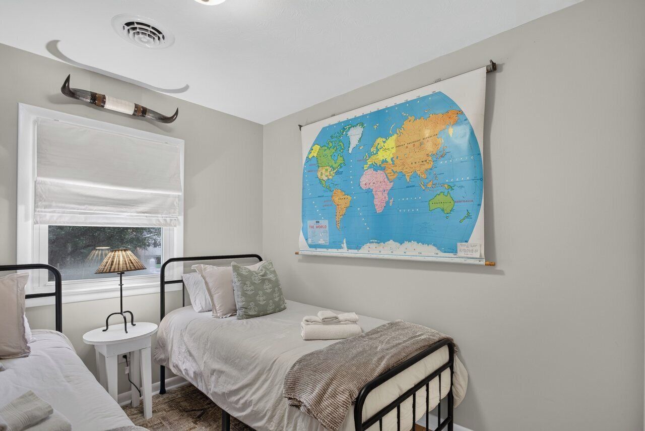 A bedroom with a bed, a small bedside table with a lamp, a window with blinds, and a world map on the wall.