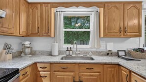 A kitchen with wooden cabinets, a granite countertop, a stainless steel sink, and a window with a view of greenery.