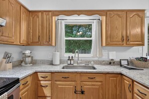 A kitchen with wooden cabinets, a granite countertop, a stainless steel sink, and a window with a view of greenery.