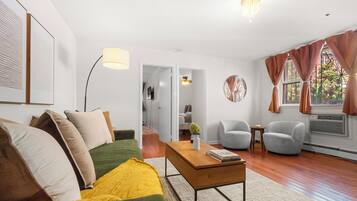 A modern living room with a green sofa, a wooden coffee table, and a grey armchair. There is a large window with curtains, a ceiling fan, and a mounted TV.