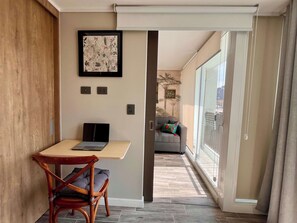 A modern living room with a wooden floor, a gray sofa, a small table with a laptop, and a large window with a view of the outside.