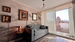 A living room with a grey sofa, a small table, and a large window with a view of buildings. - Modern Ñuñoa Apt - Projector & Free Parking (Ñuñoa)
