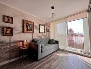 A living room with a grey sofa, a small table, and a large window with a view of buildings.
