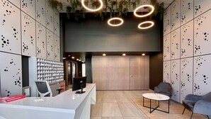 A reception area with a white reception desk, a computer, a seating area with a round table, and a wall with circular light fixtures and hanging plants. - Modern Ñuñoa Apt - Projector & Free Parking (Ñuñoa)