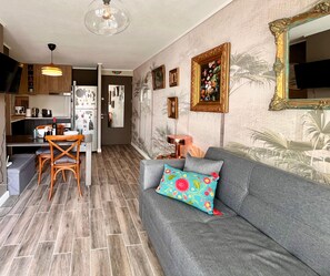 Un espacio de estar moderno con un sofá gris, un cojín colorido, una zona de comedor con mesa y sillas, y una cocina con refrigerador y armarios.