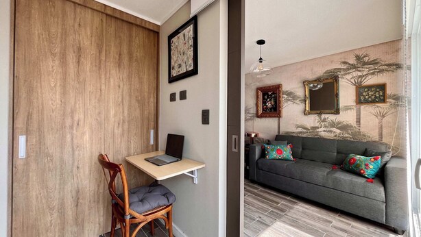 A room with a wooden sliding door, a desk with a laptop, a grey sofa with colorful cushions, and a patterned wall. - Modern Ñuñoa Apt - Projector & Free Parking (Ñuñoa)