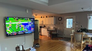 Ein geräumiges Wohnzimmer mit einem großen Flachbildfernseher an der Wand, einem Holzessttisch mit Stühlen, einer Küchenzeile mit weißen Schränken und einem kleinen Teppich.