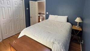 Ein Schlafzimmer mit einem Bett, einer Holzkiste, einem Deckenventilator, einem Nachttisch mit Lampe und einem Fenster mit Tageslicht.