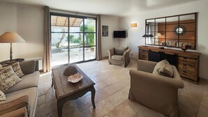 Un salon comprenant un canapé, un fauteuil et une table basse en bois. Un téléviseur est fixé au mur et une fenêtre offre une vue sur la verdure à l’extérieur.