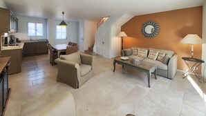 Un vaste espace de vie comprenant un canapé, une table basse et une table à manger avec des chaises. On aperçoit une cuisine à l’arrière-plan, avec une fenêtre qui laisse entrer la lumière naturelle.