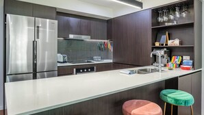 Una cocina moderna con un refrigerador de acero inoxidable, un microondas y un tostador. Hay un lavamanos con grifo, una encimera con tabla de cortar y un taburete pequeño.