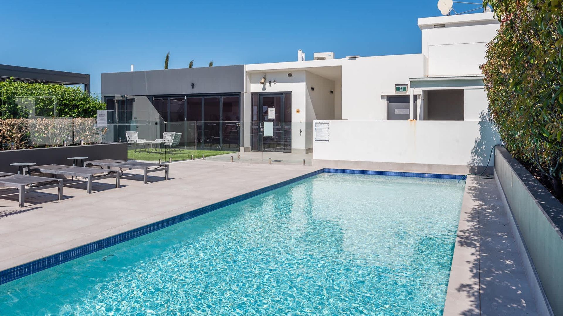 Ein moderner Außenpool mit glasklarer Wasseroberfläche, umgeben von einer Terrassenfläche mit Sitzgelegenheiten und einem im Hintergrund liegenden Gebäude mit Glasfassade.