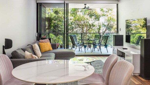 Una sala moderna con un sofá, una mesa de centro y un televisor. Hay un balcón con una mesa y sillas, y vistas a árboles.