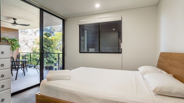 Un dormitorio con cama, ventana con persianas, ventilador de techo y balcón con sillas y plantas.