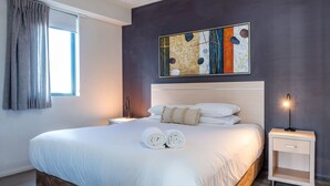 A neatly made bed with white linens and two rolled towels, a modern abstract painting on the wall, and a bedside lamp on the right.