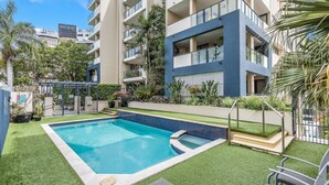 Une piscine entourée de gazon synthétique, avec un immeuble à plusieurs étages en arrière-plan.