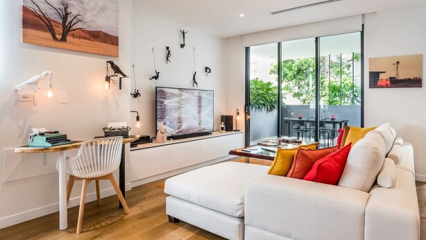 A modern living room with a white sofa, a desk with a typewriter, and a large window offering a view of greenery outside. - East Brisbane Stay - Balcony, Parking & City Access (East Brisbane)