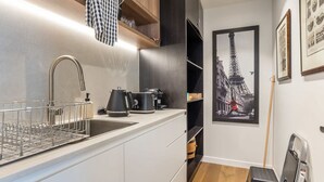A compact kitchen with a modern faucet, a kettle, and a coffee maker. There is a metal dish rack and a framed picture of the Eiffel Tower on the wall. - East Brisbane Stay - Balcony, Parking & City Access (East Brisbane)