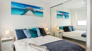 A bedroom with a bed, two bedside tables, a mirror, and artwork depicting a beach scene. - East Brisbane Stay - Balcony, Parking & City Access (East Brisbane)