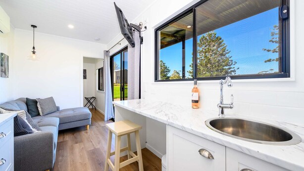 A modern kitchen with a large window, a dining area with a grey sofa, and a wooden stool.