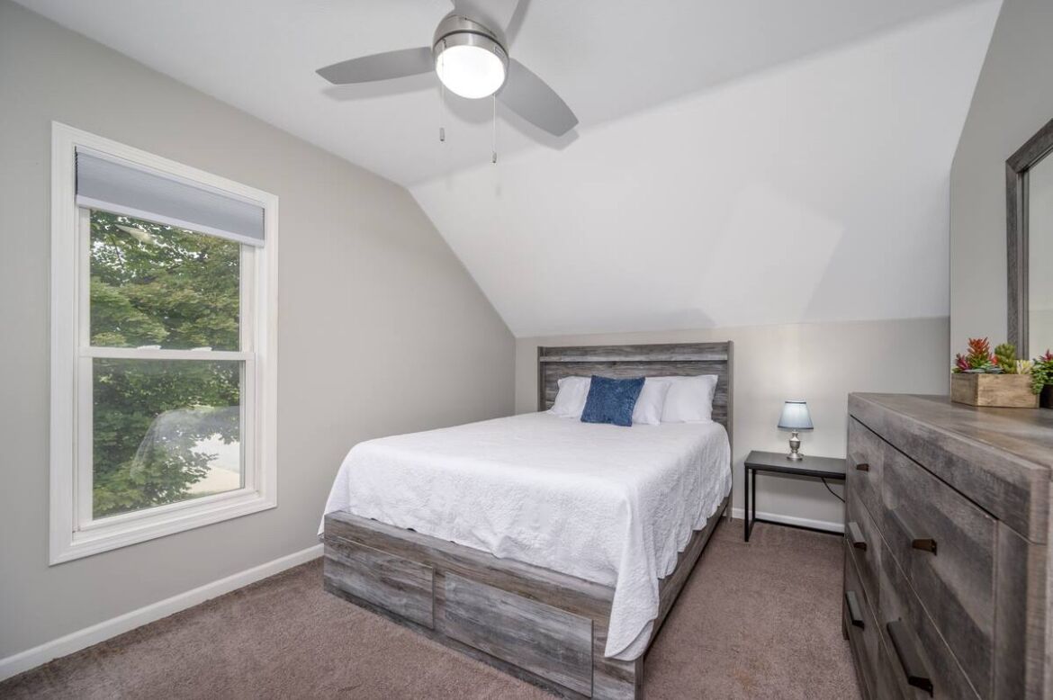 A bedroom with a bed, a nightstand, a dresser, a ceiling fan, and a window with a view of trees.