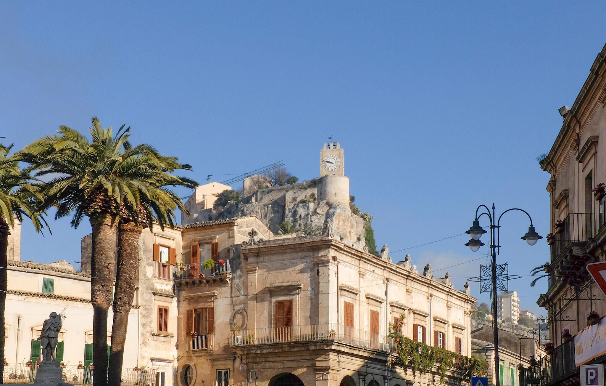 A historic building with a clock tower, palm trees, and a statue in front.