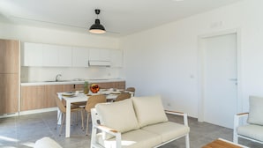 A modern living room with a sofa, chairs, a dining table with chairs, and a kitchen area with cabinets and a hanging light fixture.