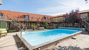 A swimming pool with a wooden deck, surrounded by a building with a tiled roof and multiple balconies. - Beautiful apartment in Svaneke with swimming pool (Svaneke)
