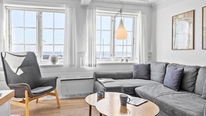 A modern living room with a grey sofa, a grey armchair, and a round wooden coffee table. There are two windows with white curtains, a hanging light fixture, and a framed picture on the wall. - Beautiful apartment in Svaneke with swimming pool (Svaneke)
