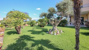 Un jardin bien entretenu, agrémenté de divers arbres et arbustes, un bâtiment doté d’un balcon et un ciel bleu dégagé.