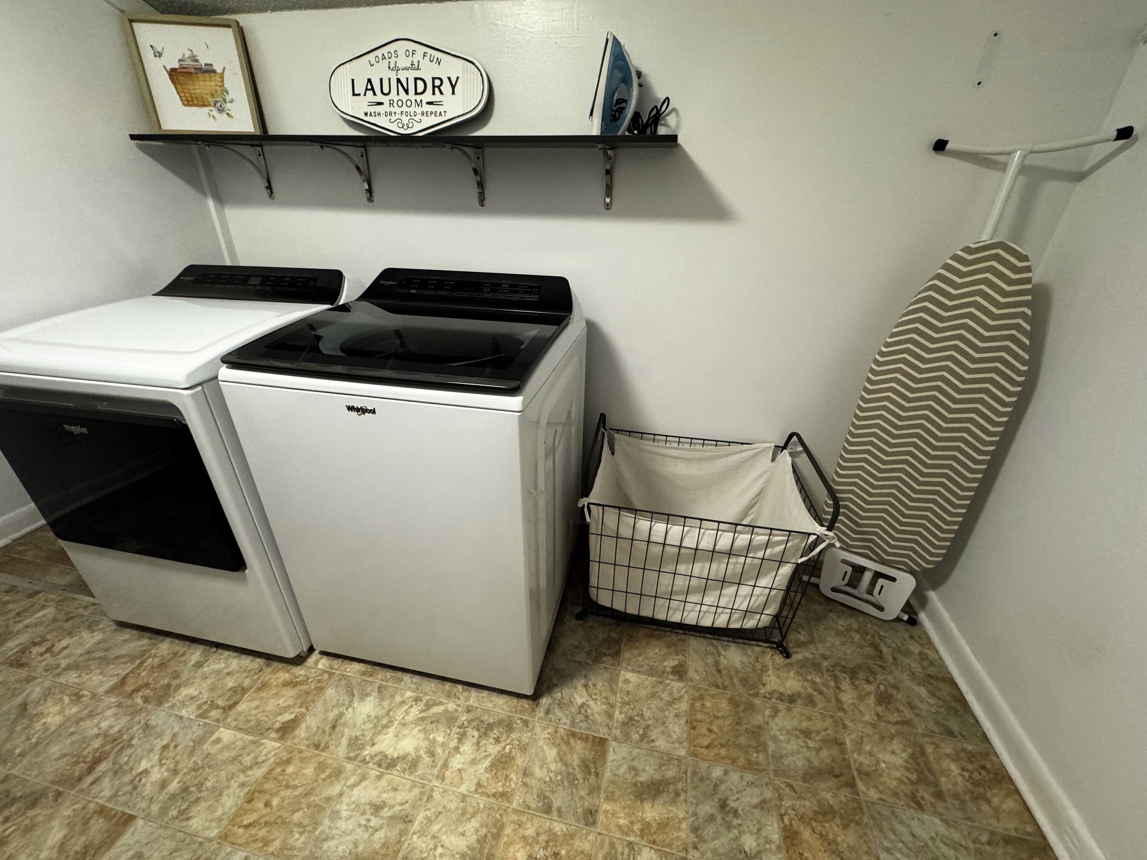 A laundry room with a Whirlpool washer and dryer, a hanging rack, a laundry basket, and a striped ironing board.