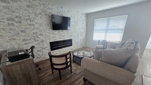 Ein modernes Wohnzimmer mit einem flachbildfernseher an einer strukturierten Wand, einem Couchtisch aus Holz, einem beigen Sofa und einem kleinen Schreibtisch mit einem Laptop.