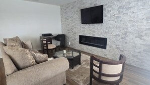 Ein modernes Wohnzimmer mit einer Wand im Naturstein-Look, einem flachbildfernseher über einem Kamin, einem Sofa, einem Couchtisch und einem Esstisch mit Stühlen.