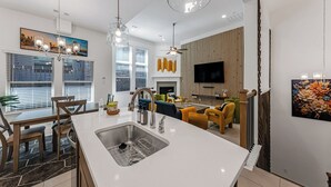 A modern kitchen with a large island, a dining area with chairs, a TV mounted on the wall, and a fireplace. - Stylish Arcade Stay EV Central (Lewisville)