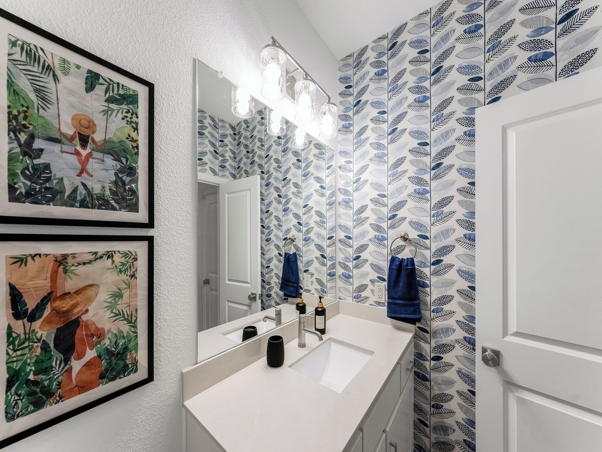 A bathroom with a white sink, a mirror with lights, and leaf-patterned wallpaper.
