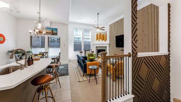 A modern kitchen with a white countertop and bar stools, a dining area with a sofa and chairs, and a staircase with a wooden railing. - Stylish Arcade Stay EV Central (Lewisville)