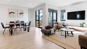Ein modernes Wohnzimmer mit einem Holzesstisch, schwarzen Stühlen, einer Couch und einem an der Wand befestigten Fernseher.