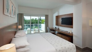 Une chambre d’hôtel avec un grand lit, un bureau avec un ordinateur, une télévision et un balcon donnant sur un espace verdoyant.
