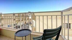 A balcony with a table and chair, overlooking a street with parked cars and buildings.