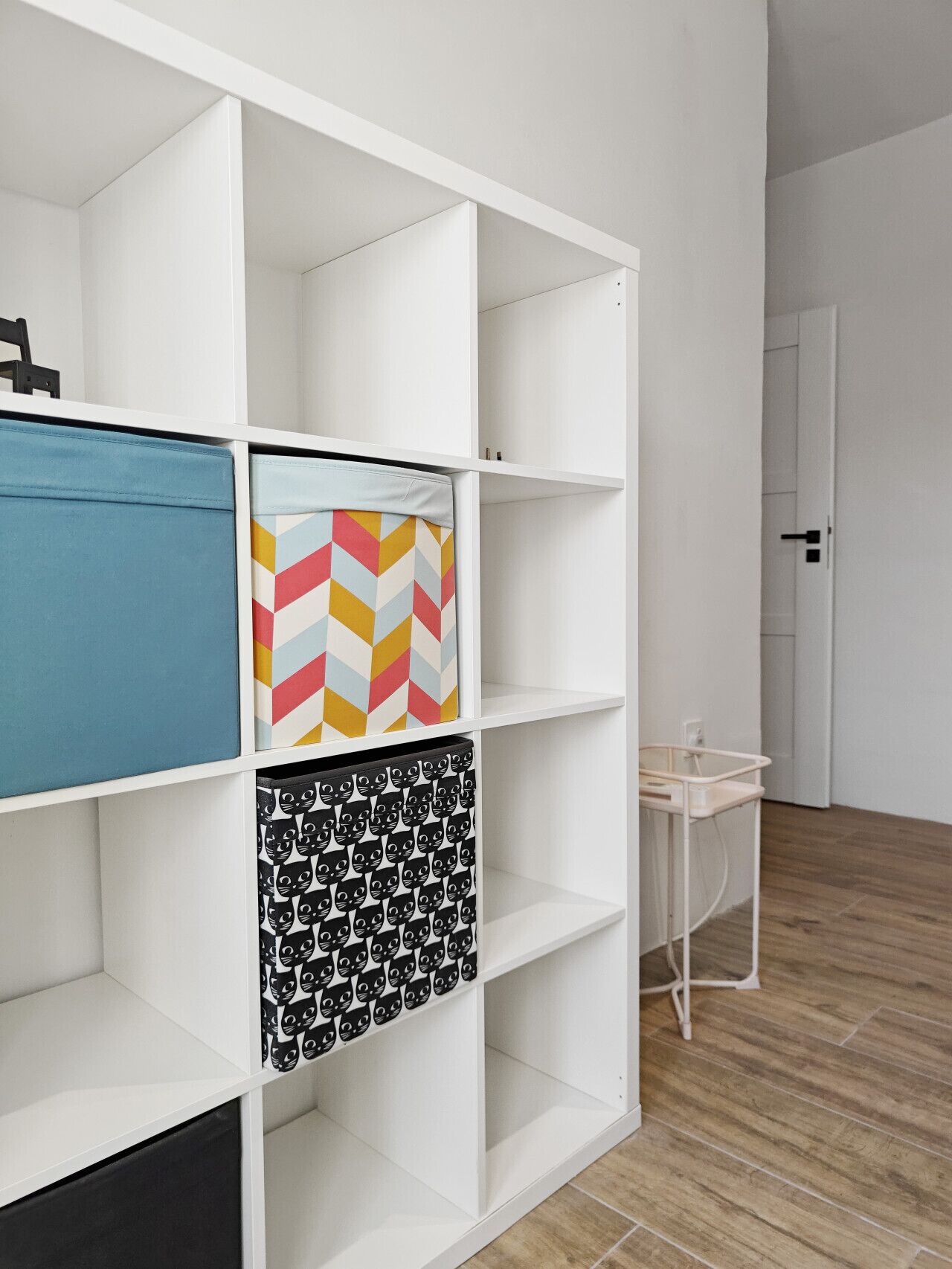Une étagère blanche avec différents bacs de rangement, un bac bleu, un coussin à motifs chevrons et un bac noir orné de motifs de chats.