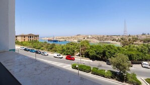 A coastal road with parked cars, a clear blue sky, and a distant view of a harbor with boats.