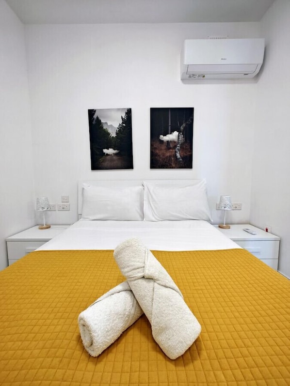 A hotel room with a bed, two nightstands, an air conditioner, and two framed pictures on the wall.