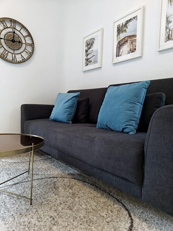 Una sala de estar moderna con un sofá gris oscuro, cojines azules, una mesa de centro redonda y un reloj de pared decorativo.
