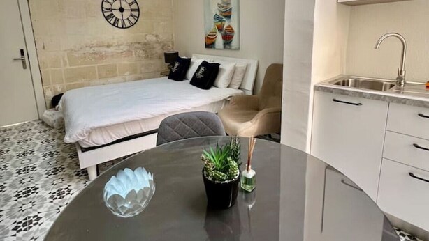 A modern bedroom with a bed, a round dining table with a plant, a wall clock, and a painting. - Cottonera Cara: Cozy Room with Terrace in the Heart of Bormla (Bormla)