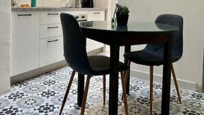 A modern kitchen with white cabinets, a black dining table, and two chairs. There is a framed plant on the wall and a tiled floor. - Cottonera Cara: Cozy Room with Terrace in the Heart of Bormla (Bormla)