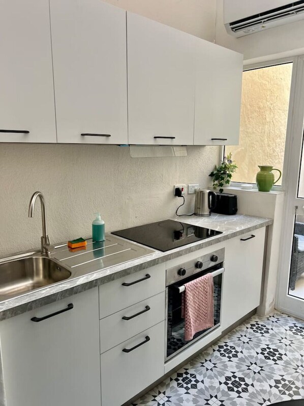 Una cocina compacta con gabinetes blancos, un lavamanos de acero inoxidable, una placa de inducción negra, un horno empotrado y un ventanal con vistas a una pared texturizada.