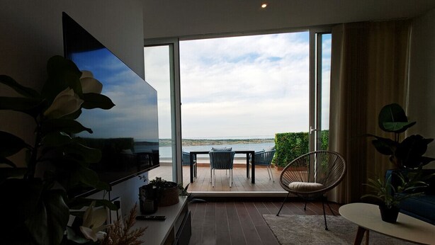 Ein modernes Wohnzimmer mit großem Fernseher, einer Couch, einem Couchtisch und Blick auf das Meer durch große Glasfenster.