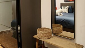 A modern hotel room with a large mirror, a bed with blue bedding, a bedside table with a woven basket, and a wooden bench.