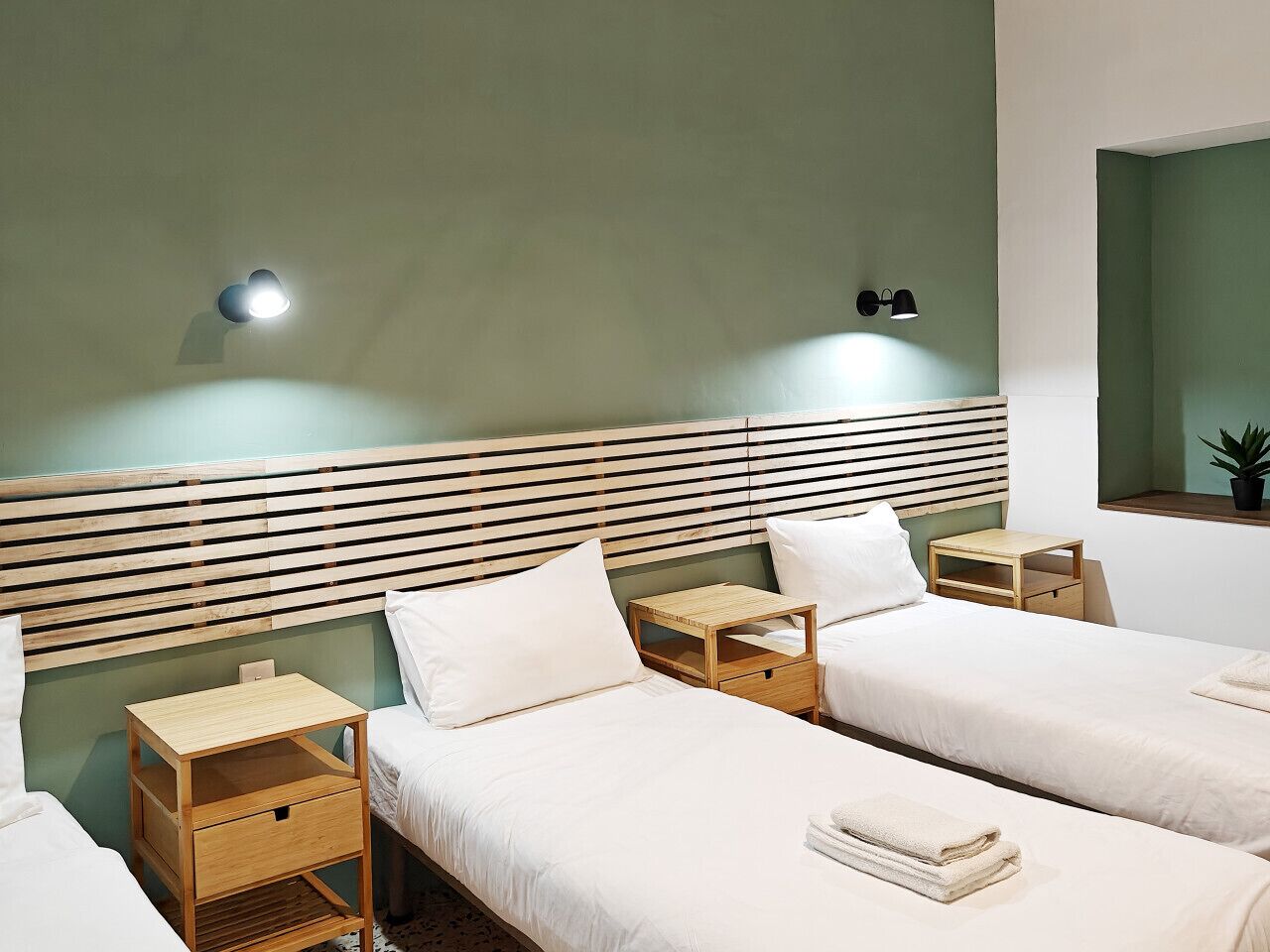 A hotel room with two beds, wooden headboards, bedside tables, and a small shelf with a plant.
